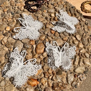 Könnte beinhalten: Vier weiße Spitzen-Schmetterlingsverzierungen auf einem Hintergrund aus kleinen braunen Steinen.
