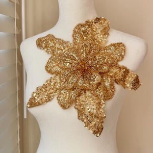 May include: A gold sequined floral applique, featuring a large flower with multiple petals and leaves. The applique is densely covered in gold sequins and beads, creating a shimmering effect. The design is displayed on a white mannequin.
