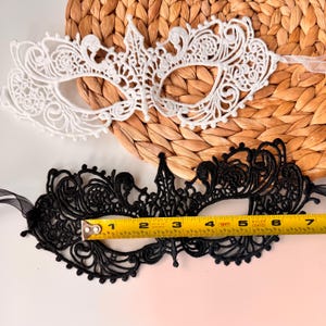 May include: Two lace masquerade masks. One is white, the other is black. The black mask is approximately 6 inches long. Both masks have intricate floral and scrollwork designs. The masks are on a woven background.