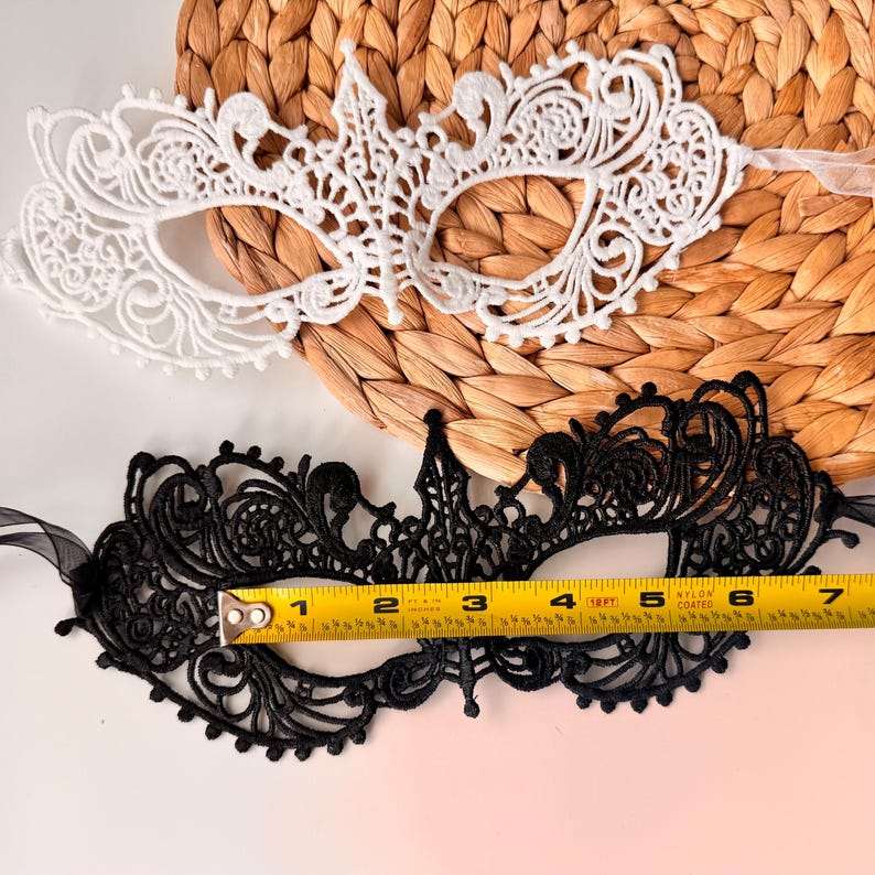 May include: Two lace masquerade masks. One is white, the other is black. The black mask is approximately 6 inches long. Both masks have intricate floral and scrollwork designs. The masks are on a woven background.