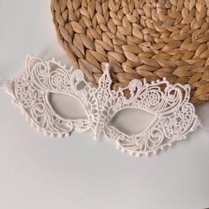 May include: A white lace masquerade mask with an intricate floral design. The mask has eye openings and a decorative crown-like element at the top. The mask is on a white surface with a woven, circular background.