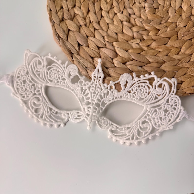 May include: A white lace masquerade mask with an intricate floral design. The mask has eye openings and a decorative crown-like element at the top. The mask is on a white surface with a woven, circular background.