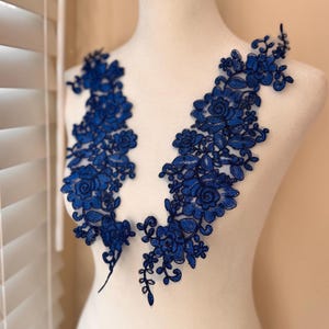 May include: Two pieces of intricate, royal blue floral lace appliques. The appliques feature detailed rose and leaf patterns, perfect for embellishing garments or craft projects. The lace is displayed on a white mannequin.
