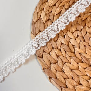 May include: A length of white floral lace trim. The delicate lace features a repeating pattern of small flowers with scalloped edges. The trim is displayed on a woven, light brown surface.