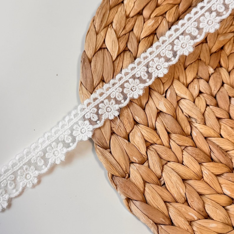 May include: A length of white floral lace trim. The delicate lace features a repeating pattern of small flowers with scalloped edges. The trim is displayed on a woven, light brown surface.