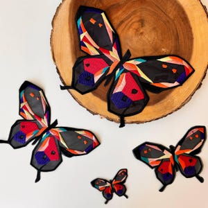 Puede incluir: Tres parches de mariposa bordados en diferentes tamaños. La mariposa más grande es negra con patrones geométricos coloridos en rojo, naranja, amarillo, azul y morado. La mariposa mediana es del mismo diseño pero más pequeña. La mariposa más pequeña es del mismo diseño pero aún más pequeña.