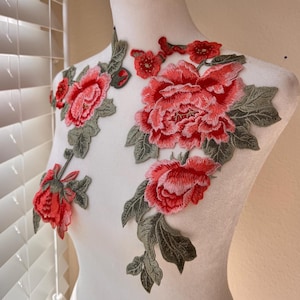 以下が含まれることがあります： 赤い花と緑の葉の刺繍のアップリケ。大きなバラと小さな花がデザインされています。アップリケは白い背景です。
