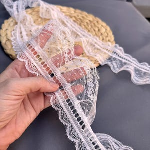May include: Close-up of white lace trim with scalloped edges and a delicate floral pattern. The lace is held in a hand, showcasing its intricate design and texture. The trim is ideal for sewing and craft projects.