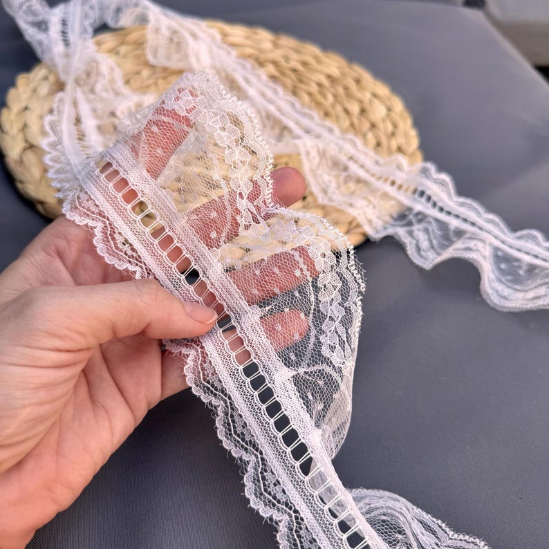 May include: Close-up of white lace trim with scalloped edges and a delicate floral pattern. The lace is held in a hand, showcasing its intricate design and texture. The trim is ideal for sewing and craft projects.
