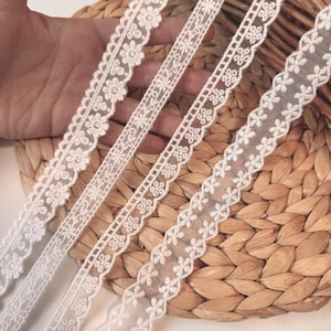 May include: Four different white lace trims with floral and geometric patterns are displayed. The lace is set against a woven, light brown background. The trims vary in width and design, showcasing delicate details.