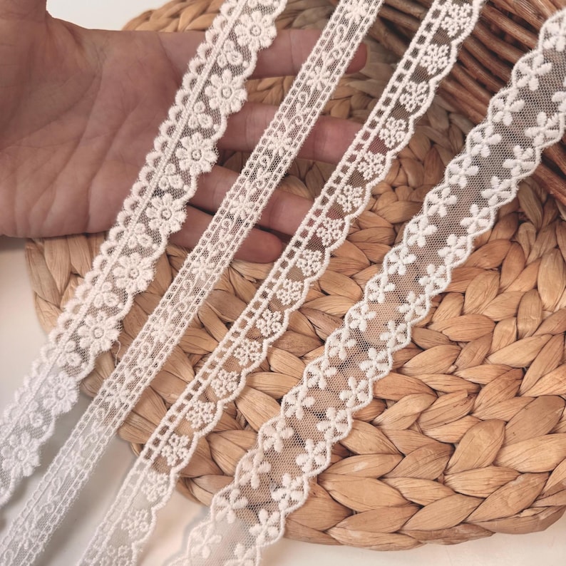 May include: Four different white lace trims with floral and geometric patterns are displayed. The lace is set against a woven, light brown background. The trims vary in width and design, showcasing delicate details.