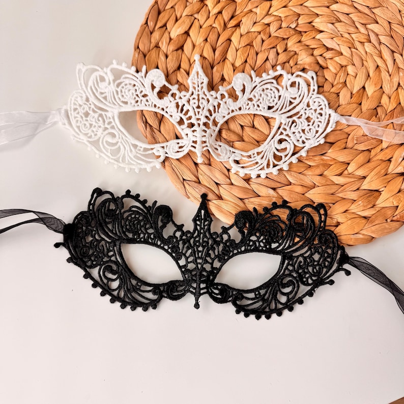 May include: Two lace masquerade masks. One is white, the other is black. Both masks have intricate floral and scrollwork designs. Each mask has sheer ribbon ties. The masks are on a woven, circular mat.