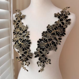 May include: Two black floral lace appliques with gold metallic outlines. The appliques are arranged symmetrically, featuring detailed rose and leaf patterns. The lace is displayed on a white mannequin, showcasing the intricate design and texture.
