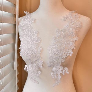 May include: Two white floral lace appliques are displayed on a mannequin. The appliques feature intricate designs of flowers and leaves, perfect for embellishing garments or crafts. The lace is delicate and detailed.