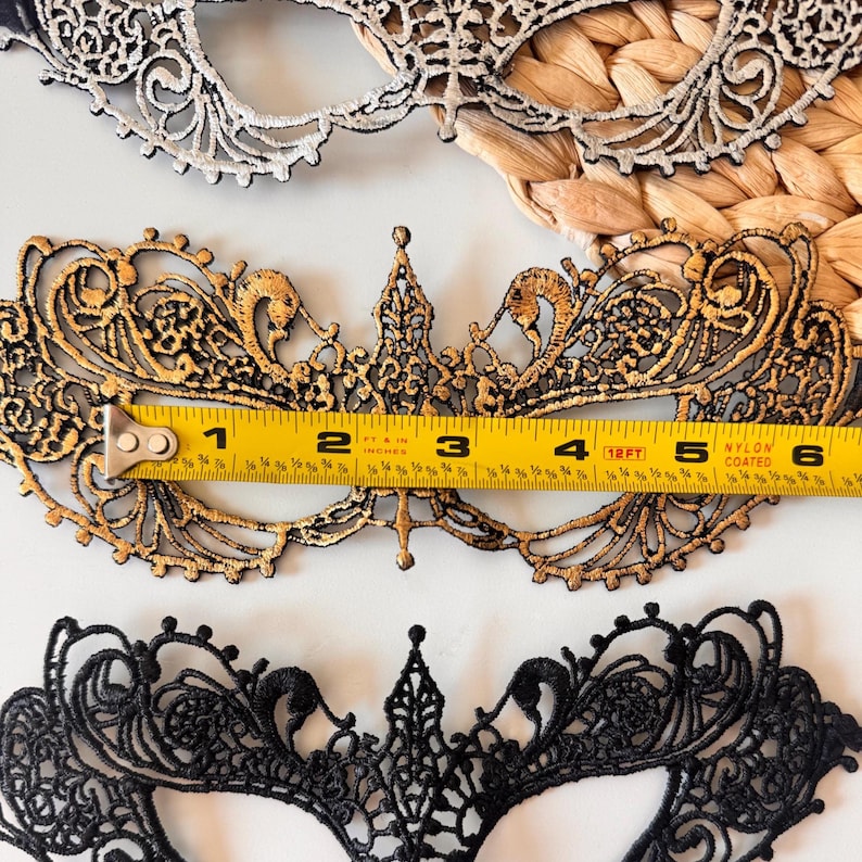 May include: Close-up of three decorative lace masks. The central mask is gold and black, measuring approximately 6 inches wide. The top mask is white and silver, and the bottom mask is black. The masks are likely for a masquerade or costume event.