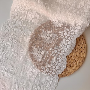 May include: Close-up of white lace fabric with floral and leaf patterns. The lace is intricately detailed with raised textures and delicate designs. The fabric is draped over a woven, circular placemat.
