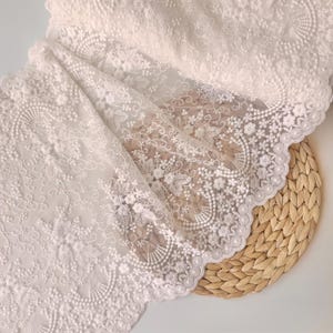 May include: Close-up of white lace fabric with intricate floral and scrollwork embroidery. The delicate lace features a scalloped edge and is draped over a woven, circular placemat. The fabric's texture and design are highlighted in the image.