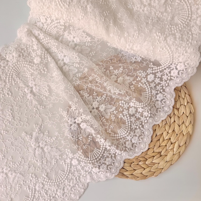 May include: Close-up of white lace fabric with intricate floral and scrollwork embroidery. The delicate lace features a scalloped edge and is draped over a woven, circular placemat. The fabric's texture and design are highlighted in the image.
