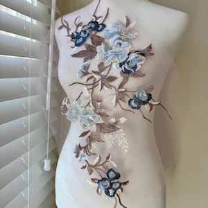Puede incluir: Aplicación floral con flores bordadas en azul y azul oscuro, tallos y hojas marrones. La aplicación está sobre una tela transparente de color claro, creando un diseño delicado y elegante. Ideal para embellecer prendas o accesorios.