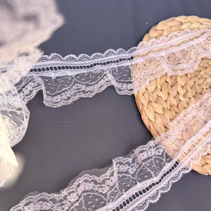 May include: Close-up of white lace trim with a scalloped edge and delicate floral patterns. The lace is layered on a dark grey surface and a woven straw mat, showcasing its intricate design and texture. Ideal for sewing and craft projects.