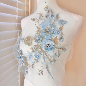 Puede incluir: Aplicación floral azul claro con detalles dorados y perlas. El diseño presenta flores y hojas bordadas, adornadas con pequeñas perlas. La aplicación está sobre una tela azul claro transparente, ideal para embellecer prendas o accesorios.
