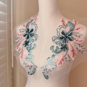 May include: Two floral appliques with pink and blue sequins and black bead accents. The appliques are on a sheer fabric background.