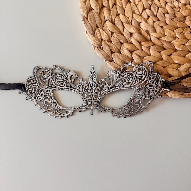 May include: Silver filigree masquerade mask with intricate lace-like detailing. The mask has eye openings and is attached to a black ribbon. The mask is a decorative accessory.