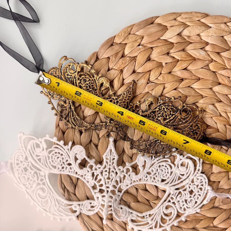 May include: Two decorative masks, one gold-toned and one white, are displayed on a woven surface. The gold mask, approximately 8 inches long, features an intricate filigree design. The white mask has a delicate lace pattern. Black ribbons are attached.