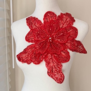 May include: A large, red sequined flower applique with multiple petals. The flower is attached to a white mannequin. The center of the flower has a cluster of small, clear beads. The petals are made of a sheer fabric.
