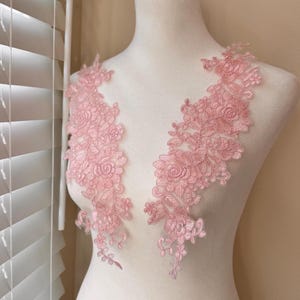 May include: Two pink floral lace appliques displayed on a mannequin. The appliques feature intricate rose and leaf designs, perfect for embellishing garments or crafts. The delicate lace is a soft, muted pink.