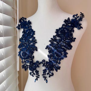 May include: Navy blue floral lace applique with intricate detailing. The applique features a symmetrical design of flowers and leaves, perfect for embellishing garments or accessories. The lace is displayed on a white mannequin.