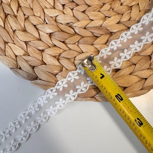 May include: White floral lace ribbon with a scalloped edge, measured with a yellow tape measure. The ribbon is approximately 5 cm wide. The background is a woven, light brown surface.