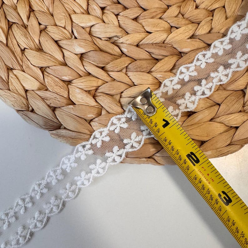 May include: White floral lace ribbon with a scalloped edge, measured with a yellow tape measure. The ribbon is approximately 5 cm wide. The background is a woven, light brown surface.