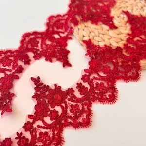 Puede incluir: Primer plano de un ribete de encaje rojo vibrante con un borde festoneado. El encaje presenta intrincados patrones florales y delicados abalorios. El ribete se muestra sobre un fondo claro, mostrando su diseño detallado.