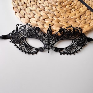 May include: Black lace masquerade mask with intricate floral and scrollwork designs. The mask has eye openings and is attached to black satin ribbons for tying. The mask is set against a neutral background.