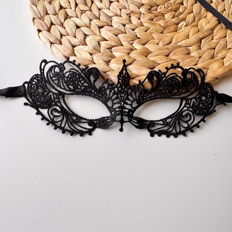 May include: Black lace masquerade mask with intricate floral and scrollwork designs. The mask has eye openings and is attached to black satin ribbons for tying. The mask is set against a neutral background.