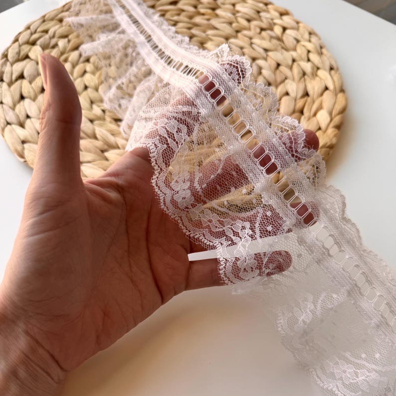 May include: Close-up of white lace trim with a scalloped edge and openwork detail. The lace, held in a hand, displays a detailed floral pattern. Suitable for sewing projects, crafts, or adding a refined touch to clothing.