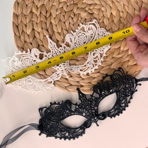 May include: Two lace masquerade masks, one white and one black, are displayed. The white mask is measured at approximately 9 inches in length. The black mask has a delicate, ornate design. Both masks are on a woven background.