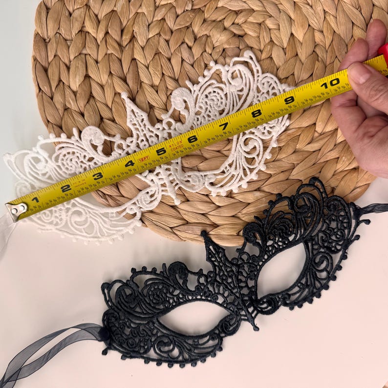 May include: Two lace masquerade masks, one white and one black, are displayed. The white mask is measured at approximately 9 inches in length. The black mask has a delicate, ornate design. Both masks are on a woven background.