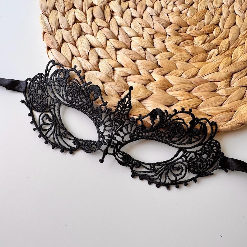 May include: Black lace masquerade mask with intricate floral and swirl patterns. The mask has a delicate, openwork design with a satin ribbon tie. The mask is set against a woven, textured background.