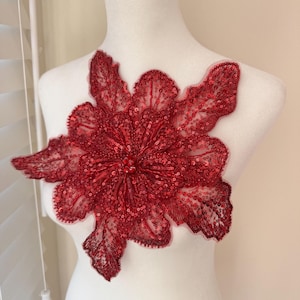 May include: A large, red floral applique with intricate sequin and bead detailing. The flower has multiple petals and a central beaded cluster. The applique is displayed on a white mannequin, showcasing its design and texture. Ideal for embellishing garments or accessories.