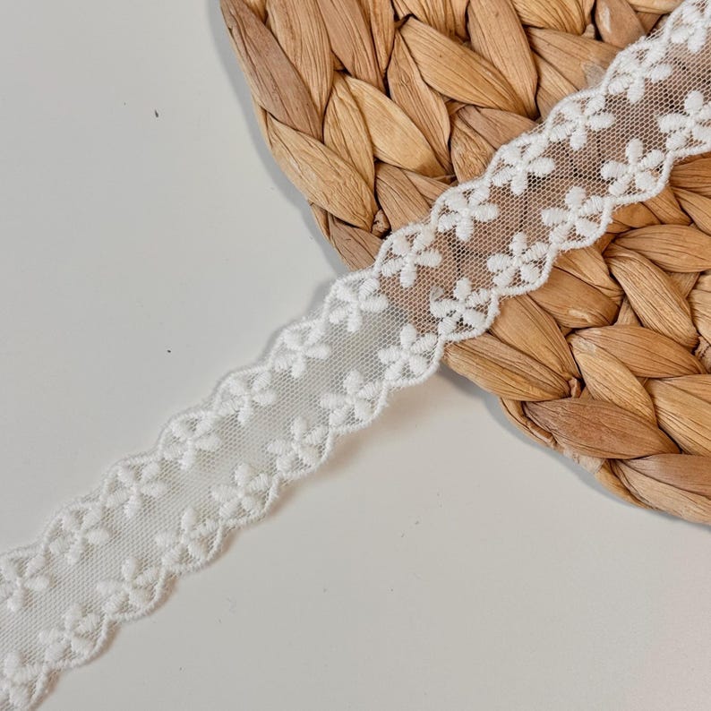 May include: White floral lace ribbon with a scalloped edge. The delicate ribbon features small, evenly spaced flower designs. The ribbon is displayed on a woven, light brown surface.