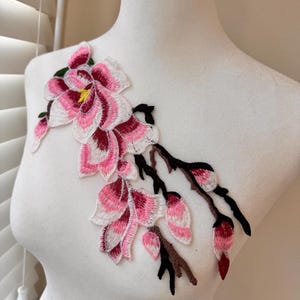 May include: Embroidered floral applique featuring pink and white petals with burgundy accents and a yellow center. The design includes brown branches and green leaves, creating a delicate, nature-inspired embellishment.