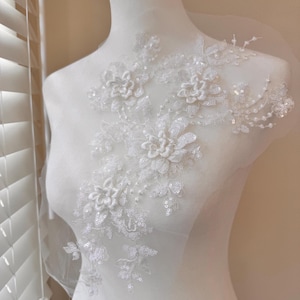 May include: White floral lace applique with 3D flower and sequin details. The applique is attached to a sheer mesh backing. The design features multiple flowers and leaves, ideal for bridal or craft projects.