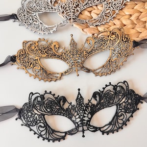 May include: Three ornate masquerade masks. The top mask is silver, the middle mask is gold, and the bottom mask is black. Each mask features intricate, openwork designs and is tied with a black ribbon. The masks are displayed on a white surface.