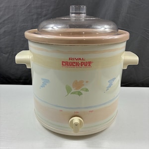 May include: A Rival Crock-Pot stoneware slow cooker with a clear glass lid. The slow cooker is off-white with pastel stripes and floral accents. It has a temperature control dial with settings for low and high.