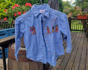 May include: A light blue button-down shirt with long sleeves. The shirt features embroidered figures of people in colorful outfits, with a lasso design. The shirt is hanging on a metal hanger outdoors.