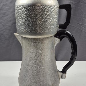 May include: Vintage silver-toned coffee percolator with a textured surface. The coffee pot has a clear glass lid and black handles. A classic kitchen appliance for brewing coffee.