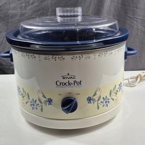 May include: Cream-colored Rival Crock-Pot slow cooker with a clear lid and blue accents. The appliance features floral designs and a control knob with settings for On, Low, and High. Includes a power cord.