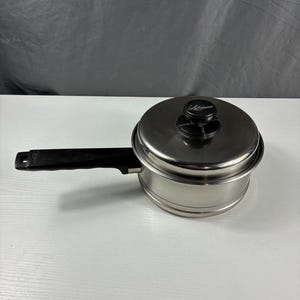 May include: Stainless steel saucepan with a black handle and lid. The lid has a black knob with the word "Lifetime" printed on it. The saucepan is sitting on a white surface.